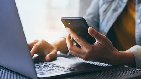 Die Hände einer Person sind zu sehen. Eine Hand liegt auf der Tastatur eines schwarzen Laptops, eine Hand hält im Vordergrund ein schwarzes Smartphone.