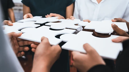 Mehrere weiße Riesen-Puzzle-Teile werden von Menschen zu einem großen Ganzen zusammengehalten.